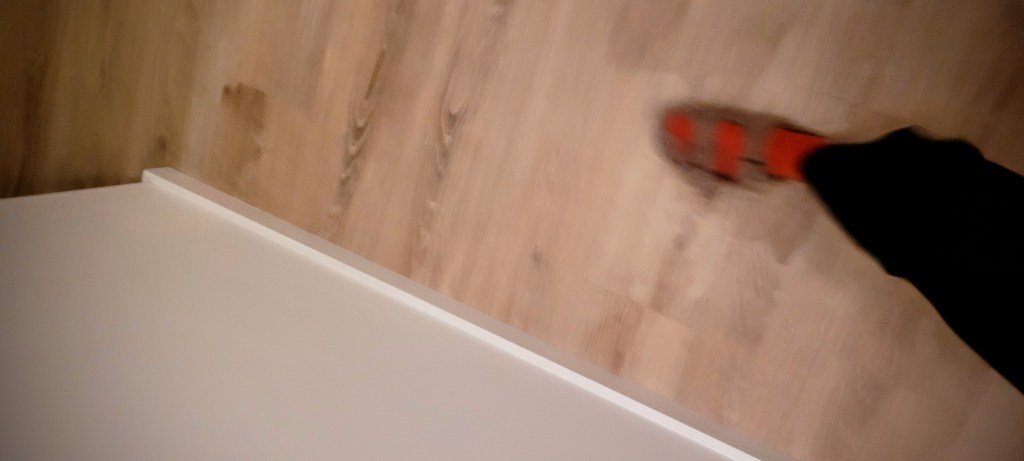 Blurry, accidental picture of a leg in black trousers. The person is wearing a red sock and a type of brown sandal. The background is a wooden floor and there is part of a wall which is white.